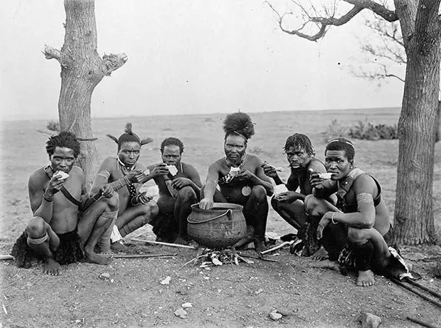Seis hombres zulúes posaron sentados en el suelo cerca de un pequeño caldero. Fotografiado entre 1920 y 1930.